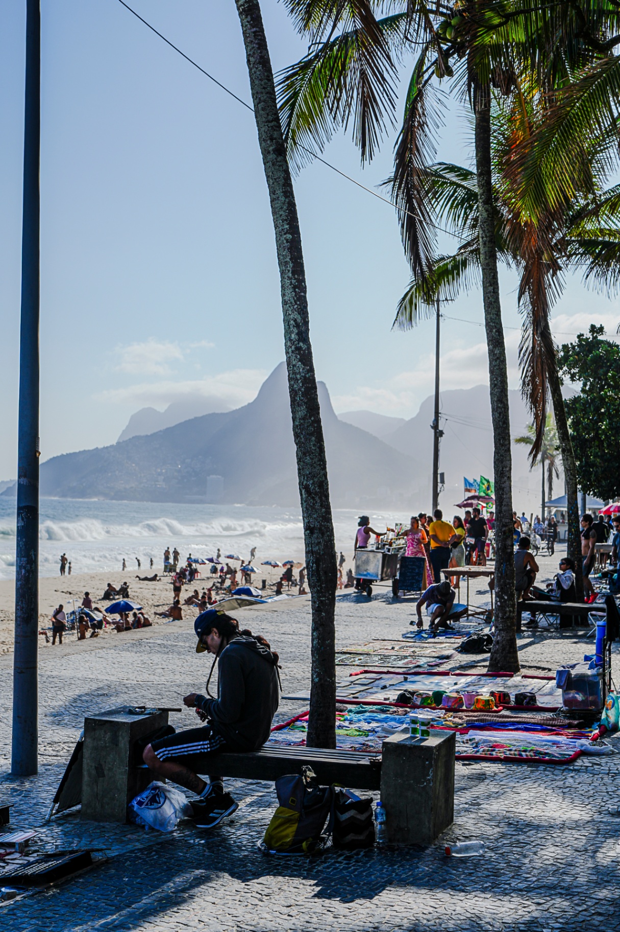 a beachside vendor seated along the ipanema promenade in rio de janeiro, with palm trees, waves, and mountains behind him.