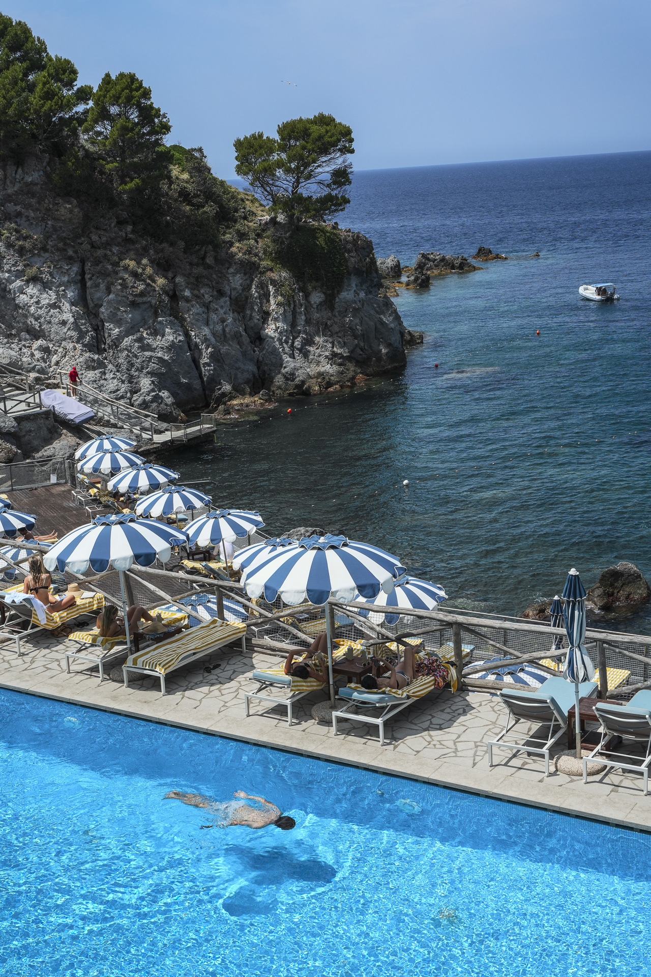 a cliffside pool on the island of ischia, italy. blue and white striped umbrellas line the deck above the tyrrhenian sea.