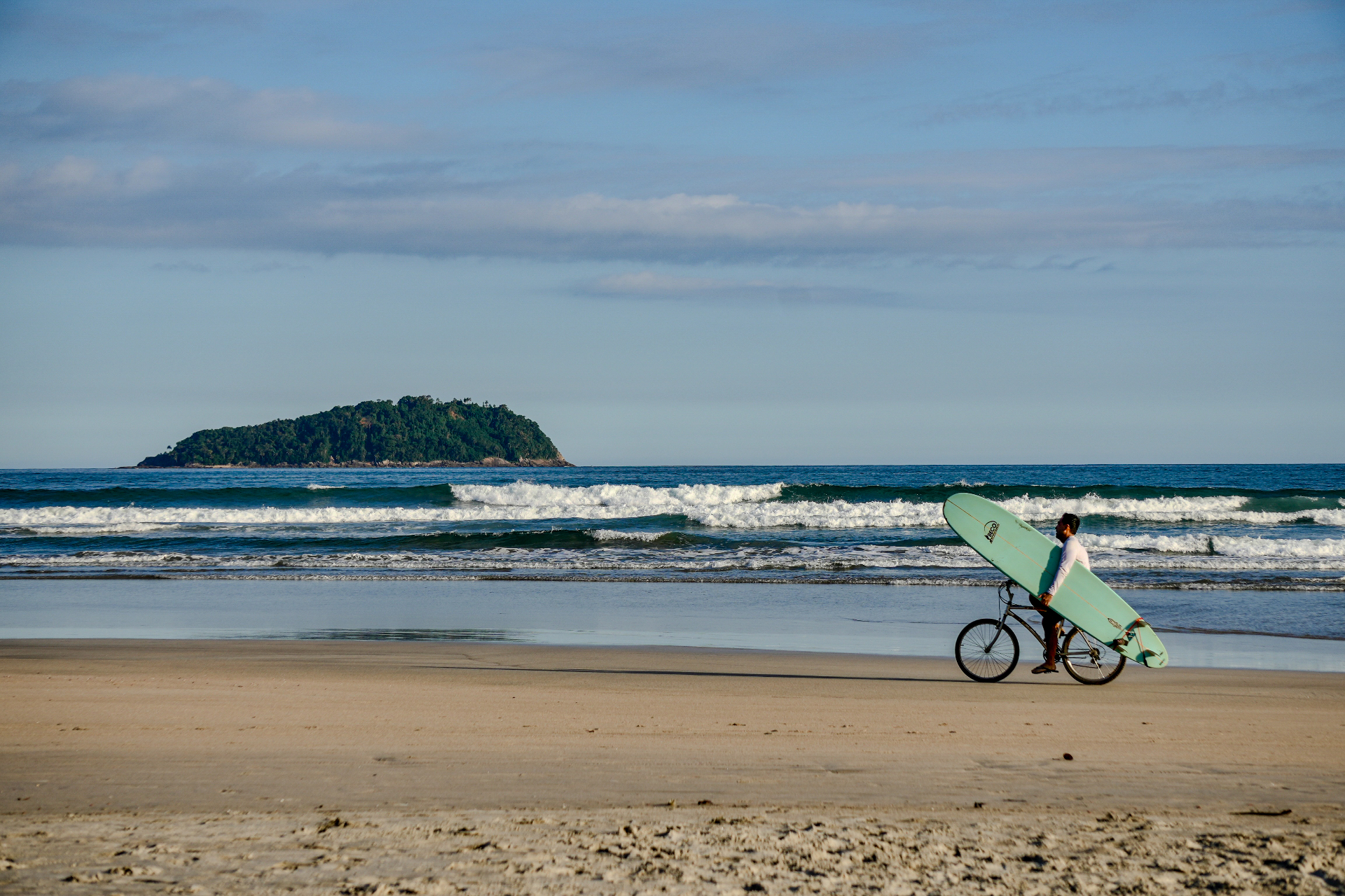 a surfer on a bicycle carrying a longboard across the sand at praia da baleia, brazil, with waves and an island in the distance.