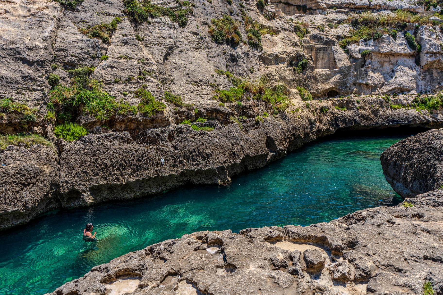 a narrow rocky inlet with clear blue-green water at wied il-għasri in gozo, malta, with a swimmer near the edge.